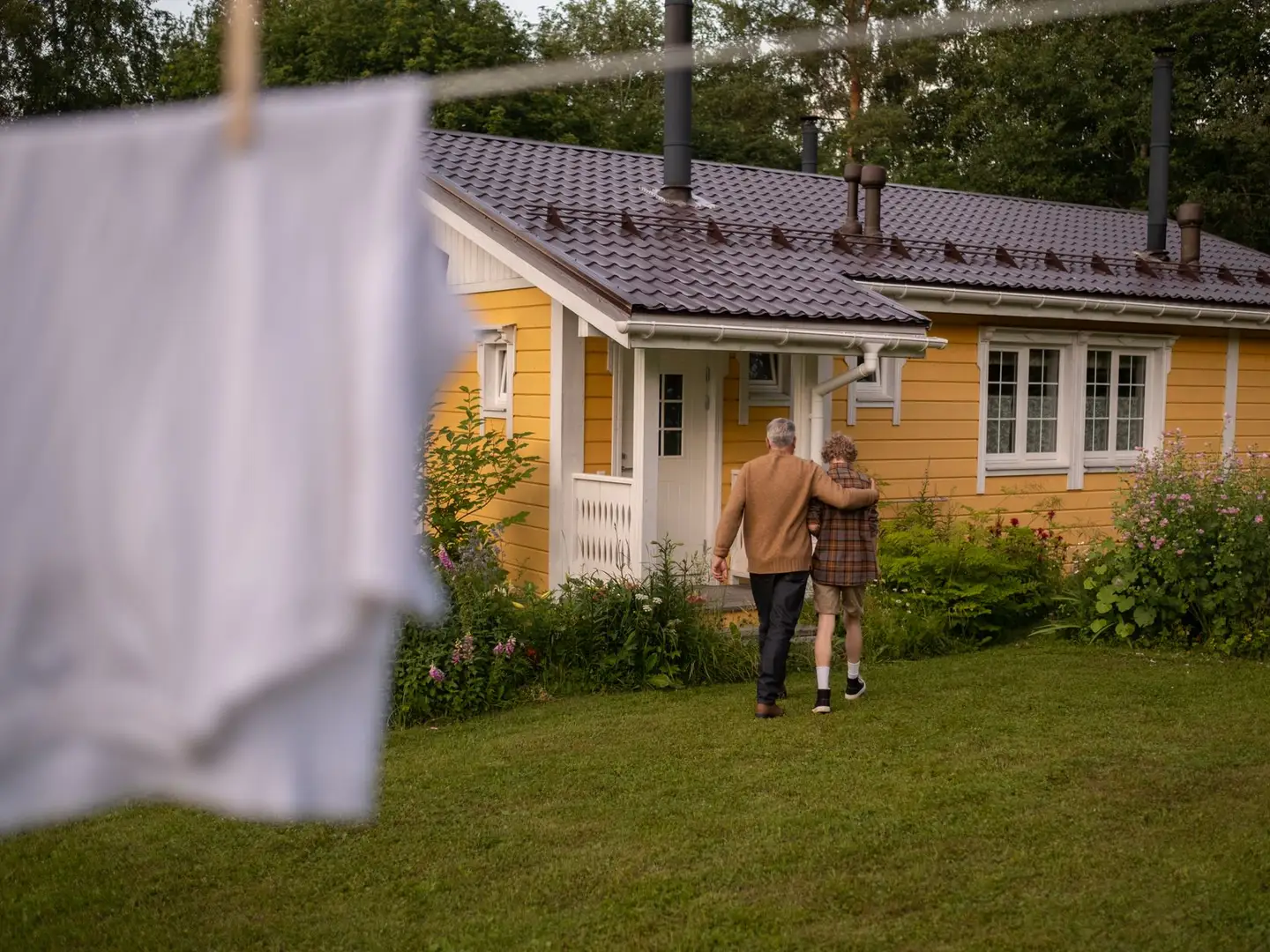 Großvater und Enkel gehen über eine Wiese auf ein gelbes Haus zu, davor hängt Wäsche an der Leine