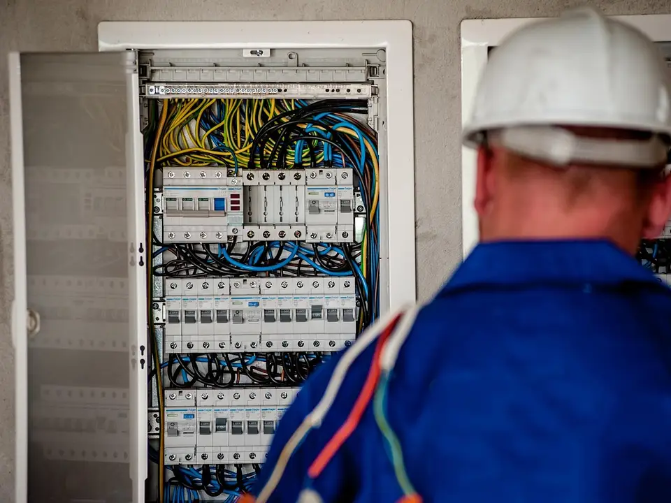 Elektriker mit Schutzhelm von hinten vor einem geöffneten Verteilerschrank mit Sicherungen