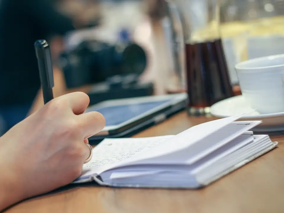 Hand mit Stift schreibt in ein aufgeschlagenes Notizbuch auf einem Tisch